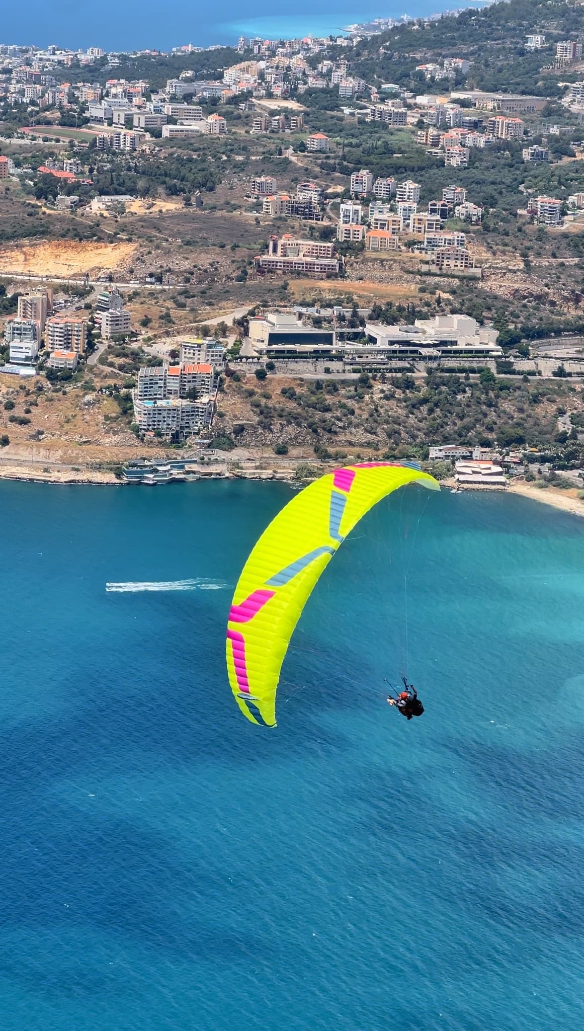 Yellow paraglider wing soaring over Jounieh Bay turquoise water and coastal city, Lebanon